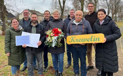 Neun Personen lächeln in die Kamera. Ein Mann hält die Urkunde in der Hand, eine Frau einen Blumenstrauß. Zwei weitere Personen halten ein Holzschild mit dem Ortsnamen "Krewelin" drauf vor sich.